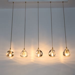 Puede incluir: Cinco l&aacute;mparas colgantes de vidrio transparente con detalles dorados cuelgan de una riel de techo blanco. Las luces est&aacute;n encendidas y emiten un brillo c&aacute;lido.