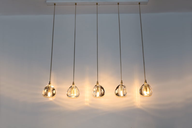 Puede incluir: Cinco l&aacute;mparas colgantes de vidrio transparente con detalles dorados cuelgan de una riel de techo blanco. Las luces est&aacute;n encendidas y emiten un brillo c&aacute;lido.