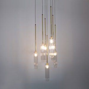 May include: A modern chandelier with multiple clear glass light fixtures hanging from a ceiling. The light fixtures are cylindrical and have a ribbed texture. The chandelier has a gold finish.