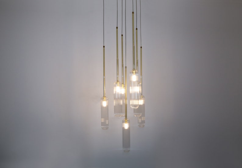 May include: A modern chandelier with multiple clear glass light fixtures hanging from a ceiling. The light fixtures are cylindrical and have a ribbed texture. The chandelier has a gold finish.