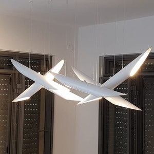 May include: A modern white ceiling light fixture with multiple arms that resemble airplane wings. The light fixture is illuminated and casts a warm glow.