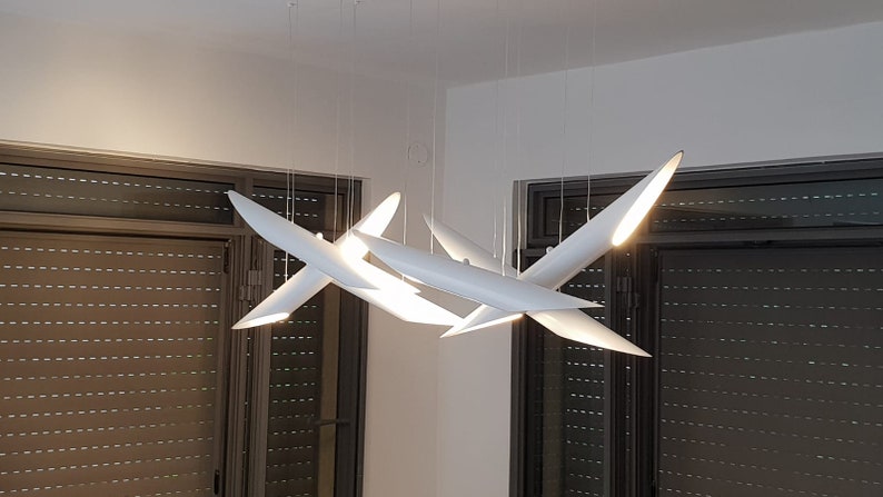 May include: A modern white ceiling light fixture with multiple arms that resemble airplane wings. The light fixture is illuminated and casts a warm glow.