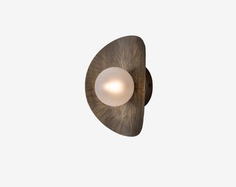 May include: A modern wall sconce with a half-moon shaped, textured, bronze-colored shade. A frosted glass globe bulb is centered within the shade. The sconce is mounted on a dark circular base.