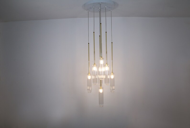 May include: A modern gold chandelier with clear glass cylinder shades. The chandelier has eight lights and is hanging from a white ceiling.