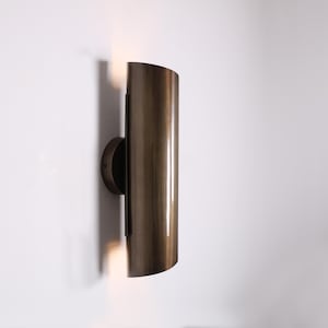Pode incluir: Arandela de parede cilíndrica de cor bronze com a parte superior curva. A arandela é montada em uma base circular e emite luz de baixo. A luminária é montada em uma parede branca.
