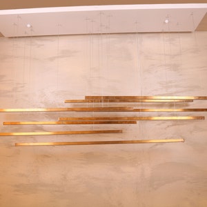 May include: A modern gold-toned chandelier with multiple horizontal bars hanging from the ceiling. The bars are suspended by thin wires and are arranged in a staggered pattern.
