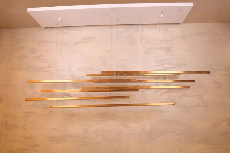 May include: A modern gold-toned chandelier with multiple horizontal bars hanging from the ceiling. The bars are suspended by thin wires and are arranged in a staggered pattern.