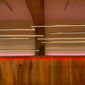 May include: A wooden wall with a pink light strip along the bottom.  There are multiple long, thin, gold-colored light fixtures hanging from the ceiling.