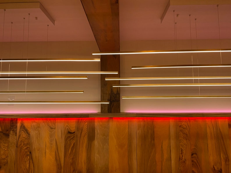May include: A wooden wall with a pink light strip along the bottom.  There are multiple long, thin, gold-colored light fixtures hanging from the ceiling.