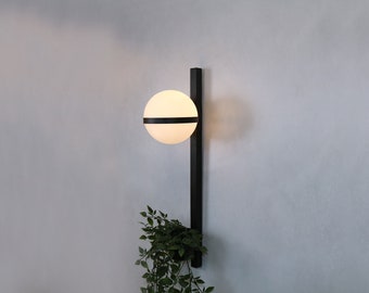 Puede incluir: Una lámpara de pared negra con un globo blanco y una banda negra en el medio. Una pequeña planta verde cuelga debajo de la luz.