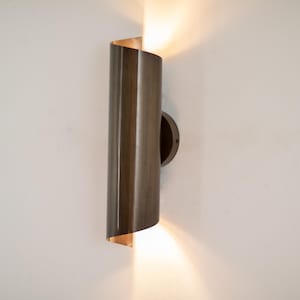 Pode incluir: Arandela de parede moderna com acabamento em bronze. A luminária cilíndrica tem a parte superior e inferior abertas, emitindo luz quente. A arandela é montada em uma base circular contra uma parede branca.