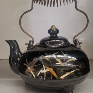 Peut inclure: Théière en céramique noire avec un motif floral décoratif blanc et doré. Elle est dotée d'une anse en métal et d'un couvercle avec un bouton doré. La théière repose sur trois petits pieds.
