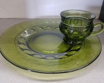 Könnte beinhalten: Ein grüner Glas-Servierteller mit erhöhtem Rand und eine kleinere grüne Glas-Tasse mit Henkel. Der Teller hat ein dekoratives Muster aus Kreisen am Rand.