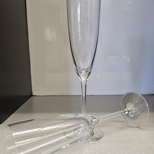 May include: Two clear champagne flutes. One stands upright, the other lies on its side. The glasses have tall, slender bowls and long stems. The background is a neutral gray and white.