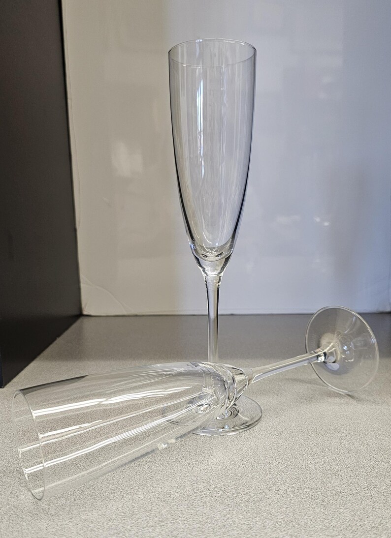 May include: Two clear champagne flutes. One stands upright, the other lies on its side. The glasses have tall, slender bowls and long stems. The background is a neutral gray and white.
