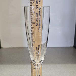 May include: A clear champagne flute with a wooden ruler placed inside. The ruler measures the height of the glass, which is approximately 9 inches tall. The flute has a slender stem and a round base.