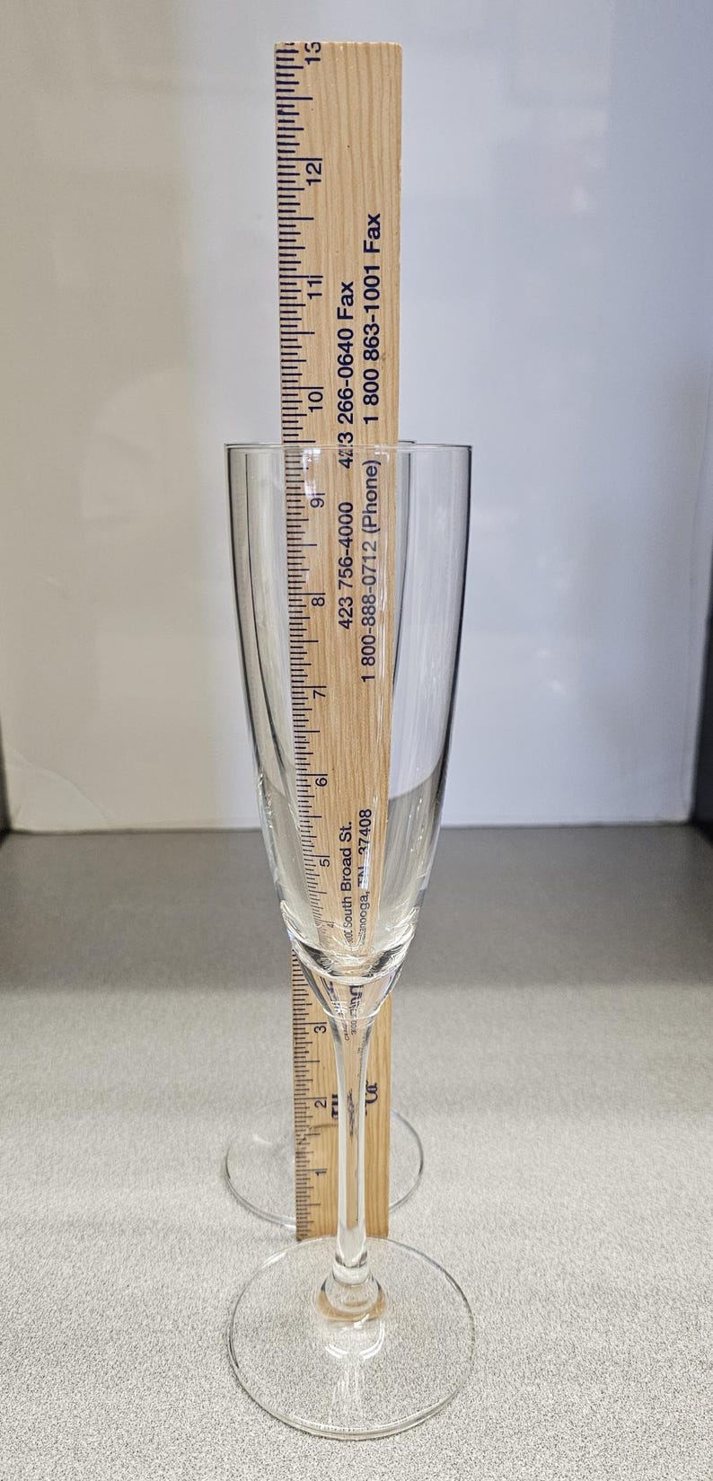 May include: A clear champagne flute with a wooden ruler placed inside. The ruler measures the height of the glass, which is approximately 9 inches tall. The flute has a slender stem and a round base.