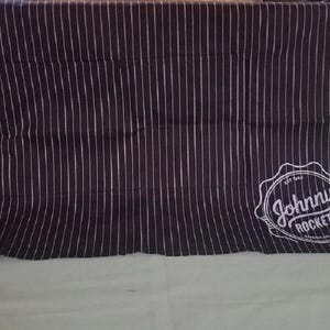 May include: A dark navy blue and white striped apron with the text "Johnny Rockets" in a white logo. The apron has a pocket and black straps for tying around the waist and neck.