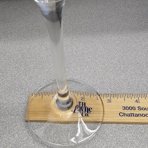 May include: A clear wine glass with a long stem, resting on a wooden ruler. The ruler shows inch measurements. The glass is positioned over the 2-inch mark. The base of the glass is visible.