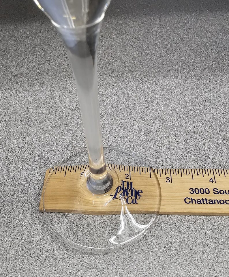 May include: A clear wine glass with a long stem, resting on a wooden ruler. The ruler shows inch measurements. The glass is positioned over the 2-inch mark. The base of the glass is visible.
