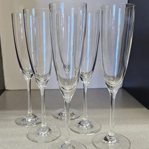 May include: A set of five clear champagne flutes. Each flute has a tall, slender bowl and a long stem, resting on a circular base. The glasses are made of transparent glass, reflecting light and creating a sparkling effect. Ideal for celebrations.