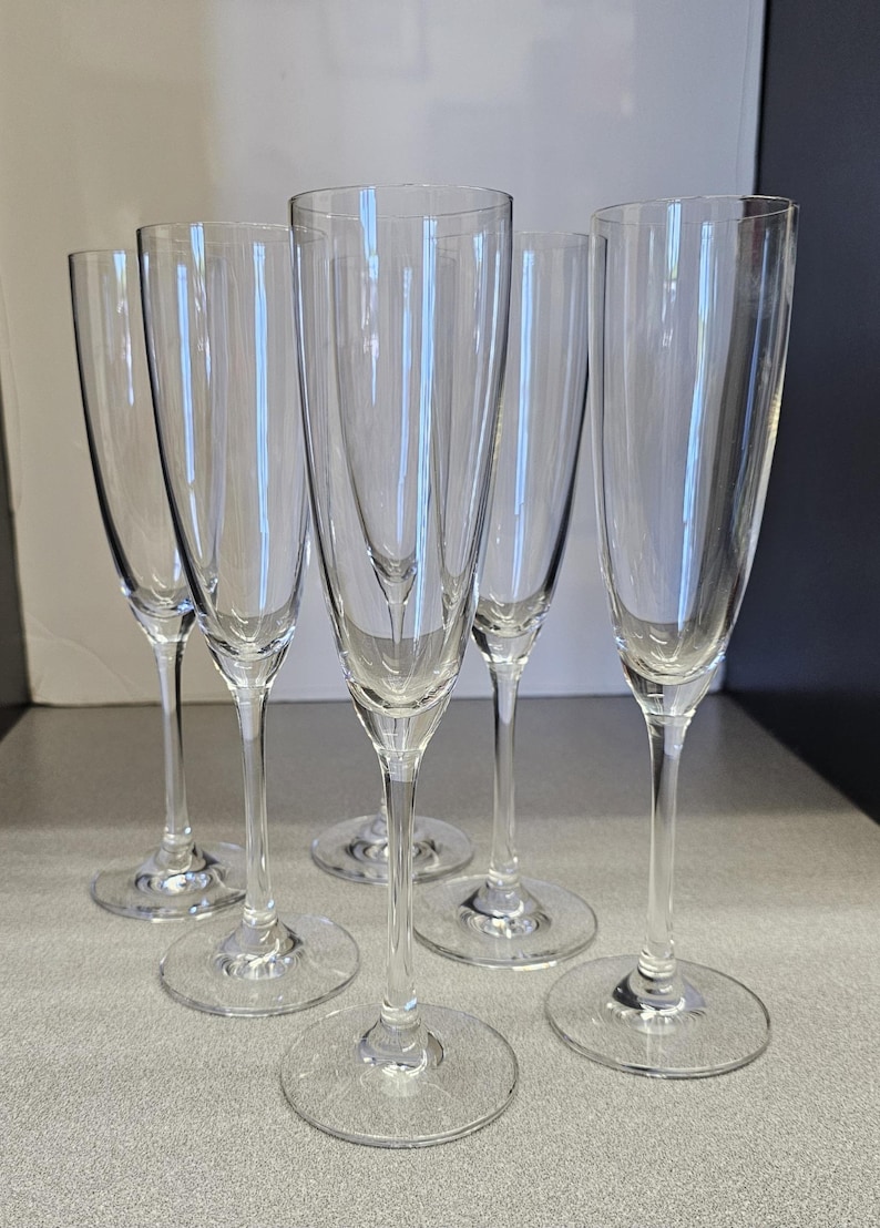 May include: A set of five clear champagne flutes. Each flute has a tall, slender bowl and a long stem, resting on a circular base. The glasses are made of transparent glass, reflecting light and creating a sparkling effect. Ideal for celebrations.