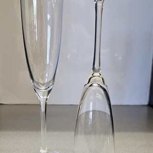 May include: Two clear champagne flutes. One is upright, the other is inverted. The flutes have a tall, slender bowl and a long stem. The base of the upright flute is round. The inverted flute's base is flat.