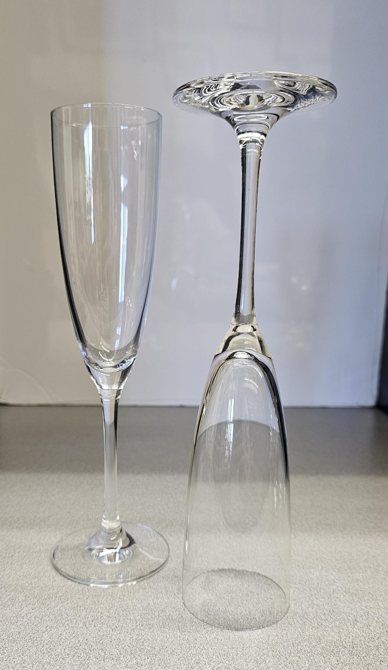 May include: Two clear champagne flutes. One is upright, the other is inverted. The flutes have a tall, slender bowl and a long stem. The base of the upright flute is round. The inverted flute's base is flat.