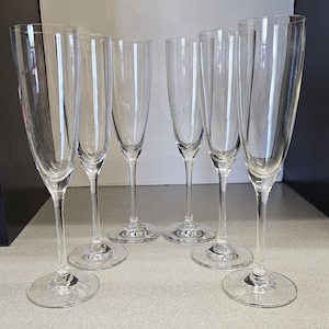 May include: Six clear champagne flutes with long stems and round bases are arranged on a light gray surface. The glasses are empty and reflect the light, showcasing their elegant design. The flutes are ideal for celebratory events.