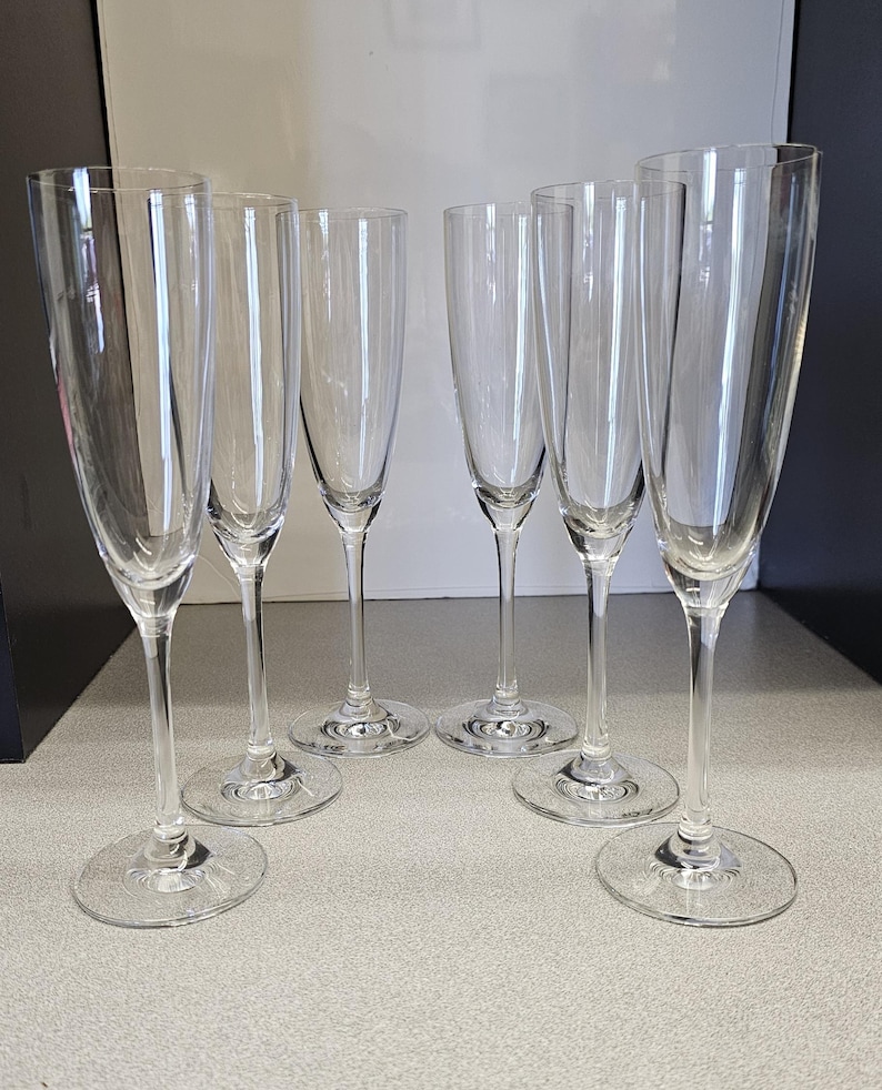 May include: Six clear champagne flutes with long stems and round bases are arranged on a light gray surface. The glasses are empty and reflect the light, showcasing their elegant design. The flutes are ideal for celebratory events.