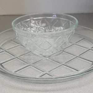 May include: A clear glass serving set consisting of a round, flat platter with a raised diamond grid pattern on the surface and a matching small, round bowl with a faceted, star-like geometric design around the exterior walls.