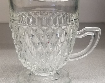 Pode incluir: Um jarro de creme de vidro transparente com um padrão de diamante e uma alça. O jarro tem uma base larga e um corpo arredondado.