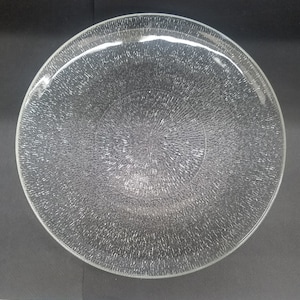 May include: A clear glass plate with a textured surface. The plate is round with a slightly raised edge. The texture appears to be a series of small, closely spaced lines radiating from the centre.