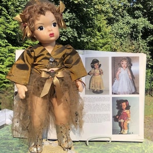 May include: A vintage doll with a brown and gold outfit, including a jacket, skirt, and boots. The doll has brown hair and is standing next to a book with images of other dolls. The doll's outfit is adorned with decorative elements.