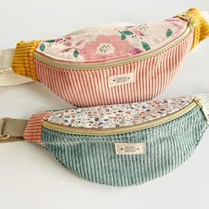 Könnte beinhalten: Zwei Gürteltaschen aus Kord mit verstellbaren Riemen und goldenen Reißverschlüssen. Die obere Tasche ist altrosa mit großem Blumenmuster und senfgelben Akzenten. Die untere Tasche ist salbeigrün mit kleinem Blümchenmuster und rosa Akzenten. Beide haben ein rechteckiges Etikett mit der Aufschrift BECKI BOOM.