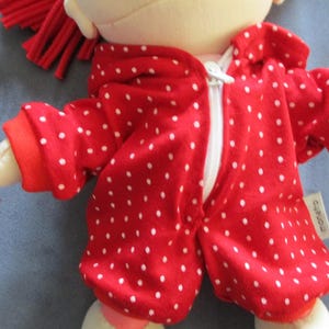 Könnte beinhalten: Eine Plüschpuppe in einem roten Overall mit weißen Punkten. Der Overall hat einen weißen Reißverschluss und rote Bündchen. Die Puppe hat rotes Haar und befindet sich auf einer blauen Oberfläche. Die Arme der Puppe sind ausgestreckt.