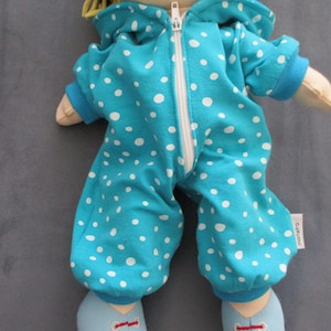 Könnte beinhalten: Puppe in einem türkisfarbenen Overall mit weißen Punkten. Der Overall hat eine Kapuze, einen weißen Reißverschluss und blaue Bündchen. Die Füße der Puppe stecken in hellblauen Schuhen mit roten Akzenten. Das Etikett lautet "monatro".