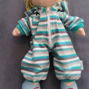 Könnte beinhalten: Handgefertigte Puppe mit einem gestreiften Overall mit Kapuze und Reißverschluss. Der Overall hat horizontale Streifen in Türkis, Grau und Weiß. Die Puppe hat blonde Garnhaare und hellblaue Schuhe.