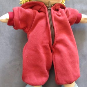Könnte beinhalten: Eine handgefertigte Puppe, die einen roten Kapuzen-Overall mit durchgehendem Reißverschluss trägt. Die Puppe hat blondes Haar und helle Haut. Der Overall hat lange Ärmel und Hosen. Die Arme der Puppe sind ausgestreckt, und die Puppe trägt hellblaue Schuhe.