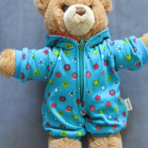 Könnte beinhalten: Ein brauner Teddybär, der einen hellblauen Strampler mit einem bunten Muster aus roten, gelben und grünen Kreisen und Sternen trägt. Der Strampler hat eine Kapuze und einen Reißverschluss vorne.