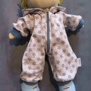 Könnte beinhalten: Eine Puppe trägt einen hellfarbigen Overall mit braunen Sternen. Der Overall hat einen durchgehenden Reißverschluss, blaue Bündchen und blaue Schuhe mit roten Schnürsenkeln. Die Puppe hat blondes Haar und einen neutralen Gesichtsausdruck.