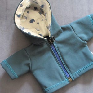 Könnte beinhalten: Türkisfarbene Kapuzenjacke mit durchgehendem Reißverschluss und kurzen Ärmeln. Die Jacke hat türkisfarbene Nähte und eine Kapuze, die mit einem cremefarbenen Stoff mit blauen Walen und nautischen Symbolen gefüttert ist.