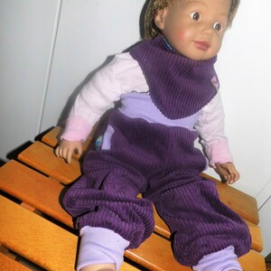 Op de afbeelding: Een paarse corduroy poppenoutfit met een bijpassende paarse corduroy slabbetje. De pop draagt ​​eronder een wit overhemd met lange mouwen. De pop heeft bruin haar en blauwe ogen.
