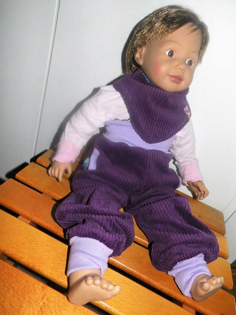 Op de afbeelding: Een paarse corduroy poppenoutfit met een bijpassende paarse corduroy slabbetje. De pop draagt ​​eronder een wit overhemd met lange mouwen. De pop heeft bruin haar en blauwe ogen.