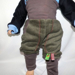 May include: Green corduroy trousers with brown trim and brown leggings underneath. The trousers have a small green and red tag attached to the right leg.