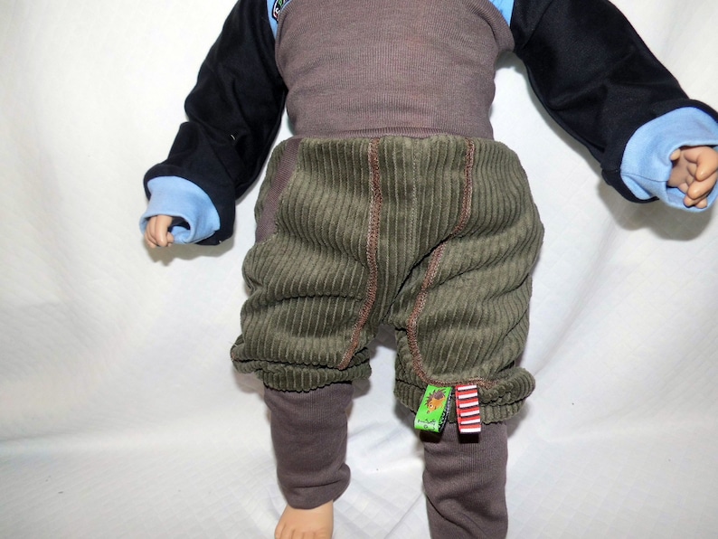 May include: Green corduroy trousers with brown trim and brown leggings underneath. The trousers have a small green and red tag attached to the right leg.