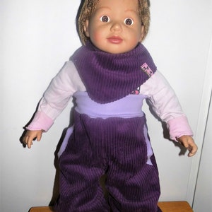 Op de afbeelding: Een paarse corduroy poppenoutfit met een bijpassende paarse corduroy slabbetje. De outfit heeft een lavendelkleurige tailleband en het slabbetje heeft een roze label met de tekst "The Baby Shop".