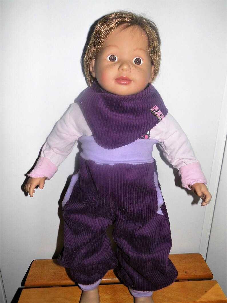 Op de afbeelding: Een paarse corduroy poppenoutfit met een bijpassende paarse corduroy slabbetje. De outfit heeft een lavendelkleurige tailleband en het slabbetje heeft een roze label met de tekst "The Baby Shop".