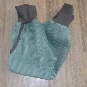 Puede incluir: Un pantal&oacute;n de pana verde con pu&ntilde;os de canal&eacute; marrones. El pantal&oacute;n est&aacute; doblado por la mitad y colocado plano sobre una superficie de madera.