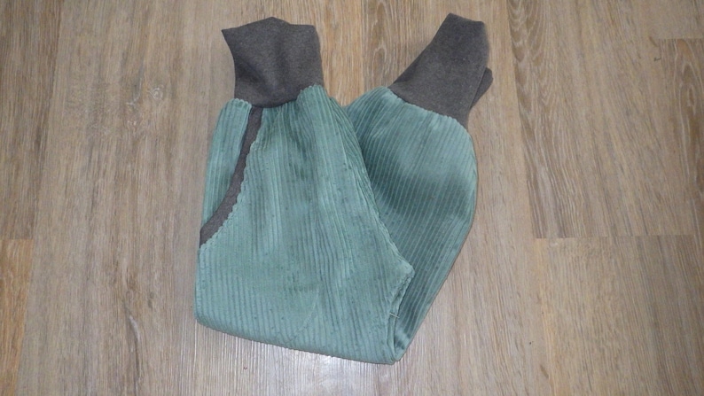 Puede incluir: Un pantal&oacute;n de pana verde con pu&ntilde;os de canal&eacute; marrones. El pantal&oacute;n est&aacute; doblado por la mitad y colocado plano sobre una superficie de madera.